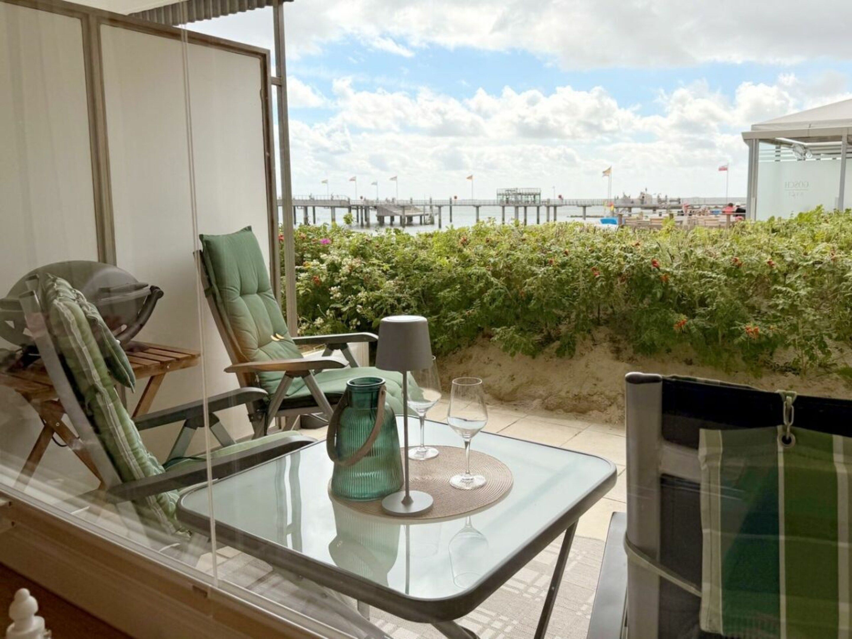 A balcony with a glass table, two chairs, a green cushion, and a glass vase.