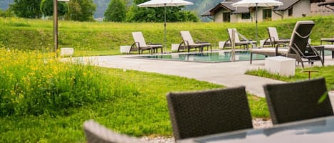 A poolside area with lounge chairs and umbrellas, surrounded by a grassy landscape and mountains in the background.