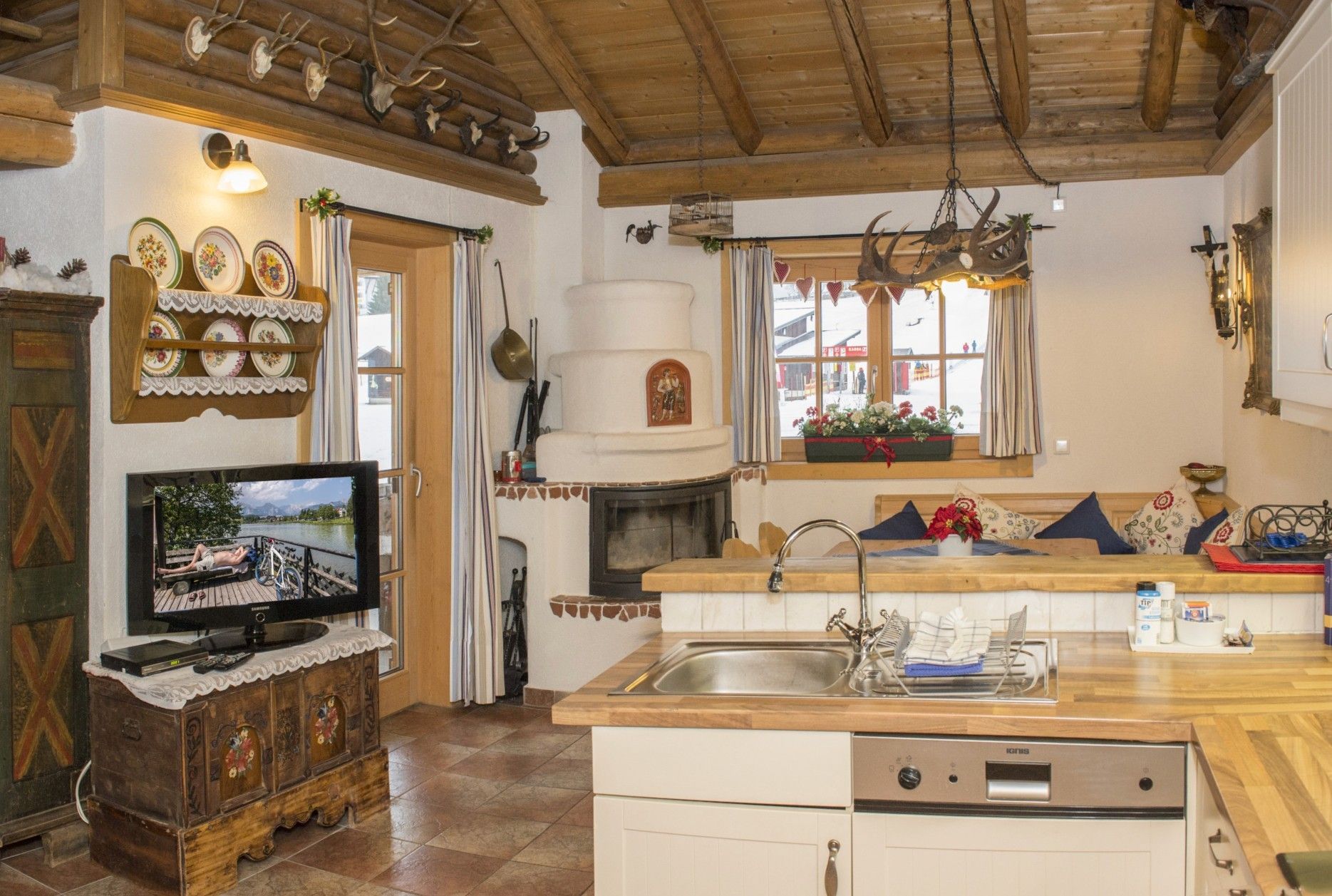 A cozy kitchen with a wooden ceiling, a television on a cabinet, a sink, and a window with a view.