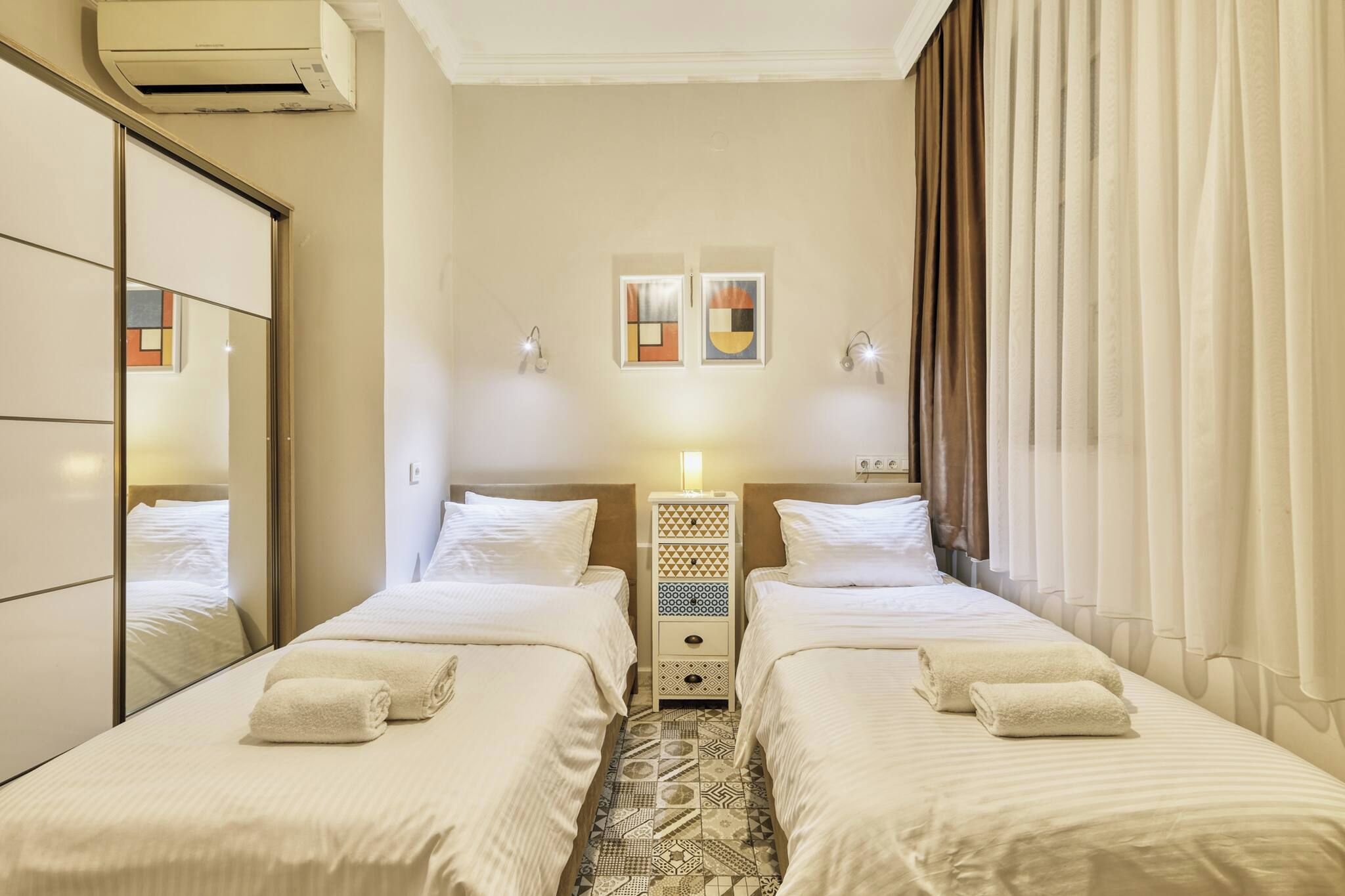 A hotel room with two beds, a bedside table with a lamp, a wall-mounted air conditioner, and a mirror.