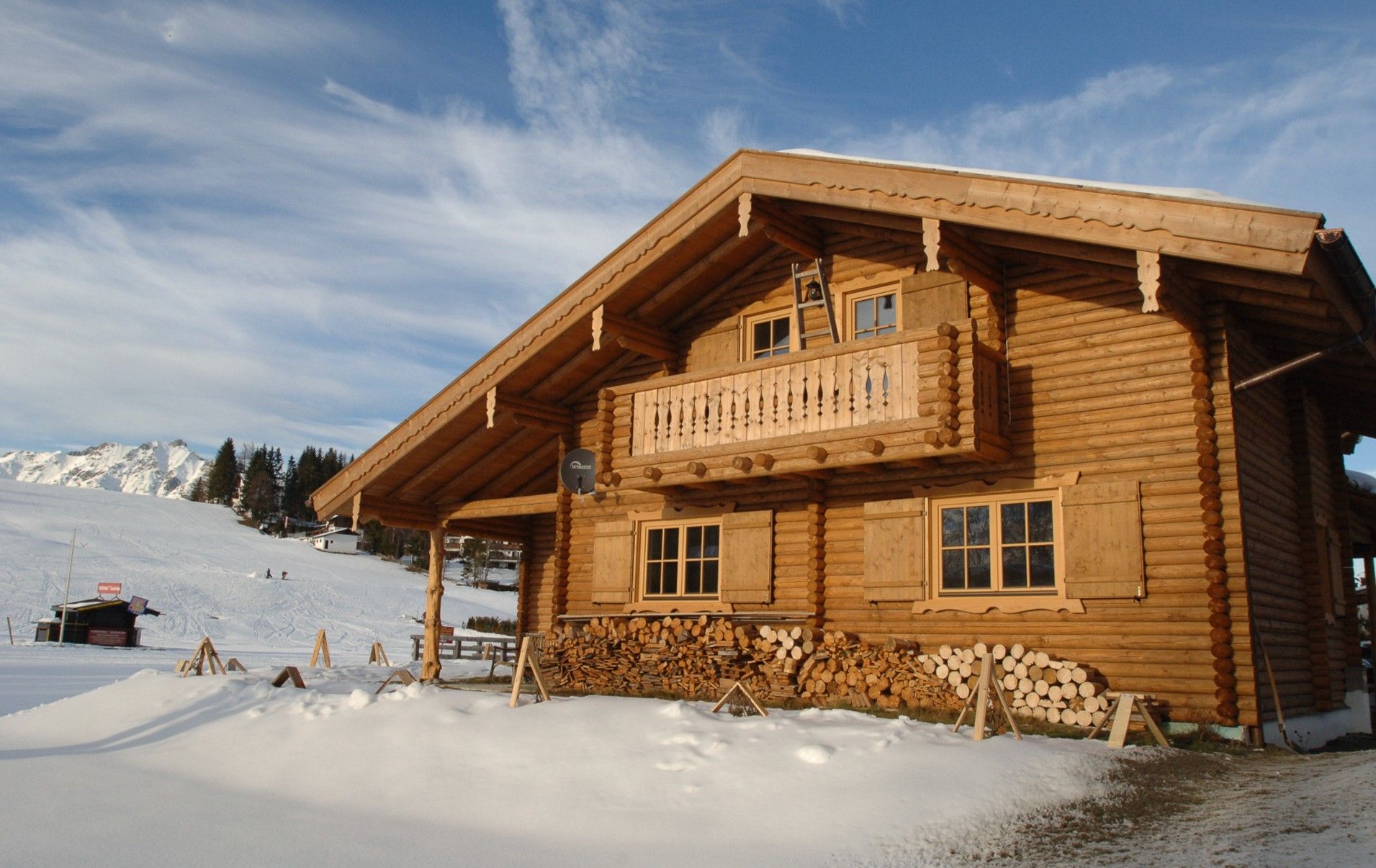 Eine Holzhütte mit Balkon, verschneiter Fläche und Bergen im Hintergrund.