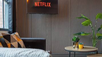 A modern living room with a television displaying 'NETFLIX,' a sofa with patterned cushions, a round coffee table with decorative items, and a large plant.