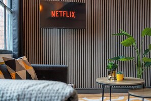 A modern living room with a television displaying 'NETFLIX,' a sofa with patterned cushions, a round coffee table with decorative items, and a large plant. - The Craftsman 3BR Apt, JQ Birmingham with Parking (Birmingham)