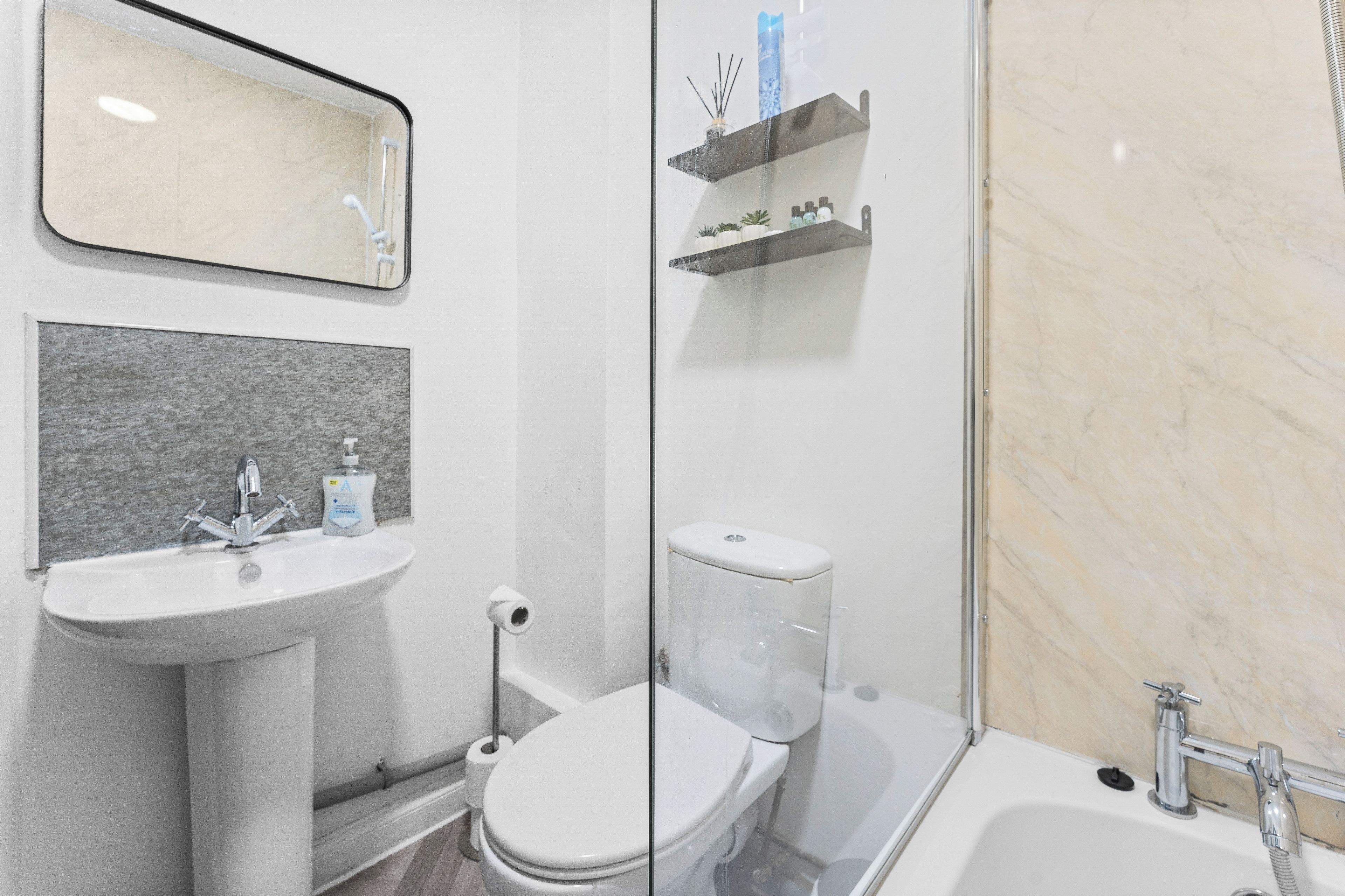 A bathroom with a white sink, toilet, and bathtub. There is a mirror above the sink and a shelf with toiletries.