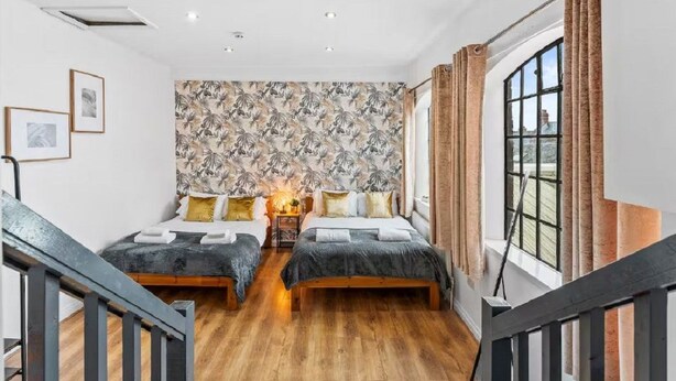Un dormitorio con dos camas, un papel tapiz estampado, una ventana con cortinas y una escalera.