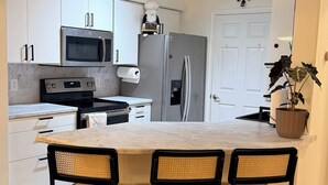 A modern kitchen with white cabinets, a stainless steel oven, a microwave, a refrigerator, and a dining area with a marble countertop and three wicker chairs. - Modern Furnished Retreat Cozy & Convenient (Castle Rock)