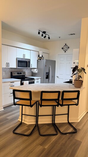 A modern kitchen with white cabinets, a stainless steel oven, a microwave, a refrigerator, and a dining area with a marble countertop and three wicker chairs. - Modern Furnished Retreat Cozy & Convenient (Castle Rock)