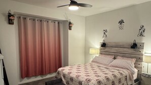 Een slaapkamer met een bed, een plafondventilator, een raam met gordijnen en een nachtkastje met een lamp.
