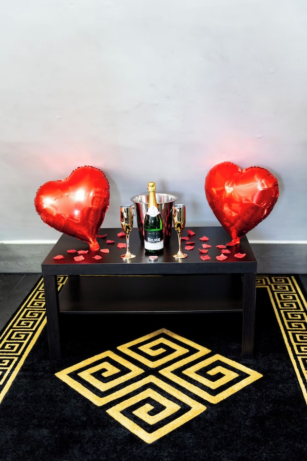A table with two heart-shaped balloons, a bottle of Belnor champagne, and two champagne flutes on a black table with a geometric patterned rug.