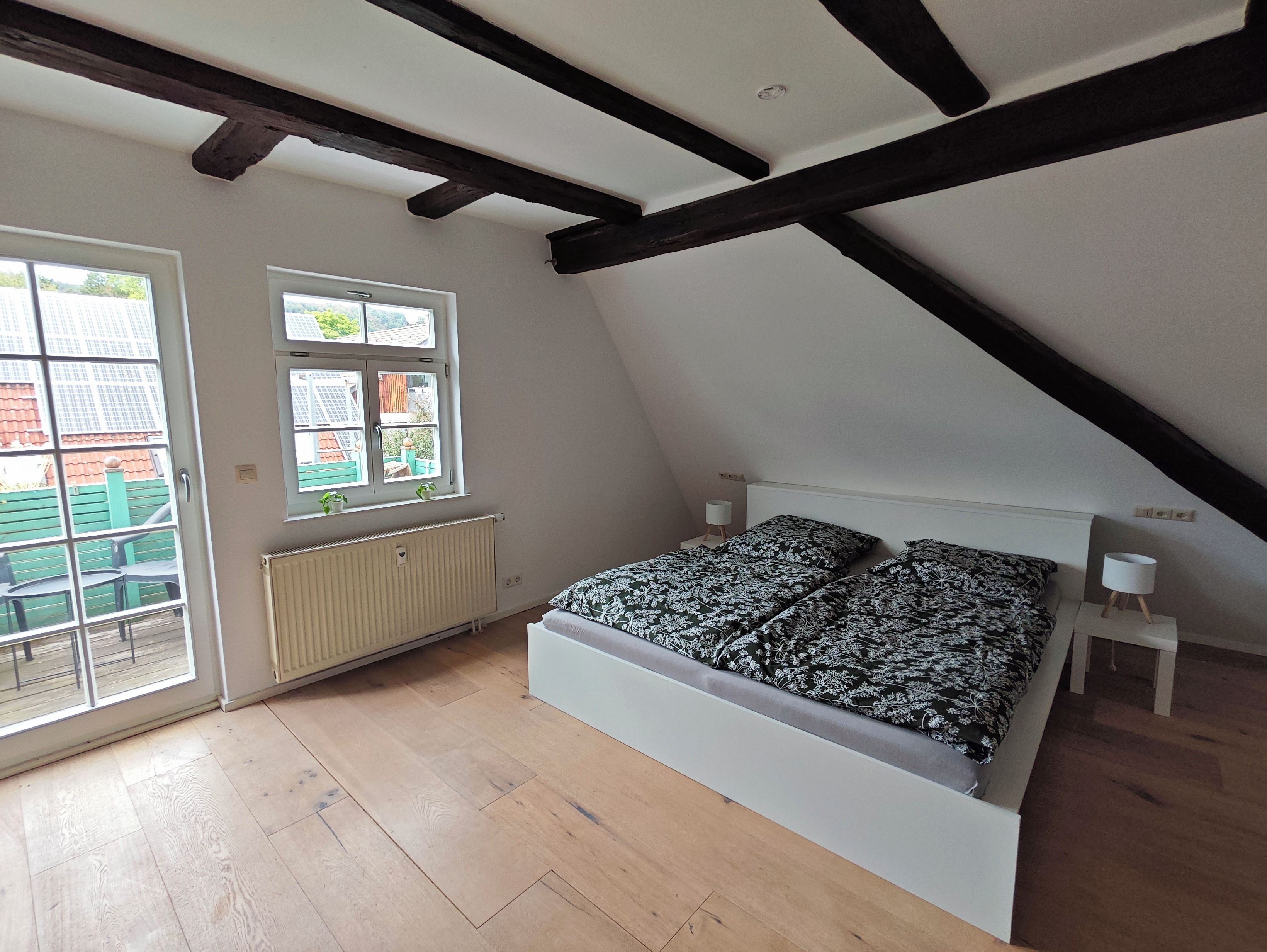Ein Schlafzimmer mit geneigter Decke, einem Bett mit gemusterten Betttextilien, einem Heizkörper und einem Fenster mit Blick auf Gebäude.