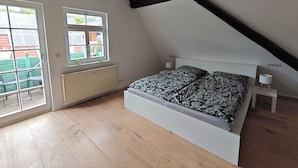 Ein Schlafzimmer mit geneigter Decke, einem Bett mit gemusterten Betttextilien, einem Heizkörper und einem Fenster mit Blick auf Gebäude.