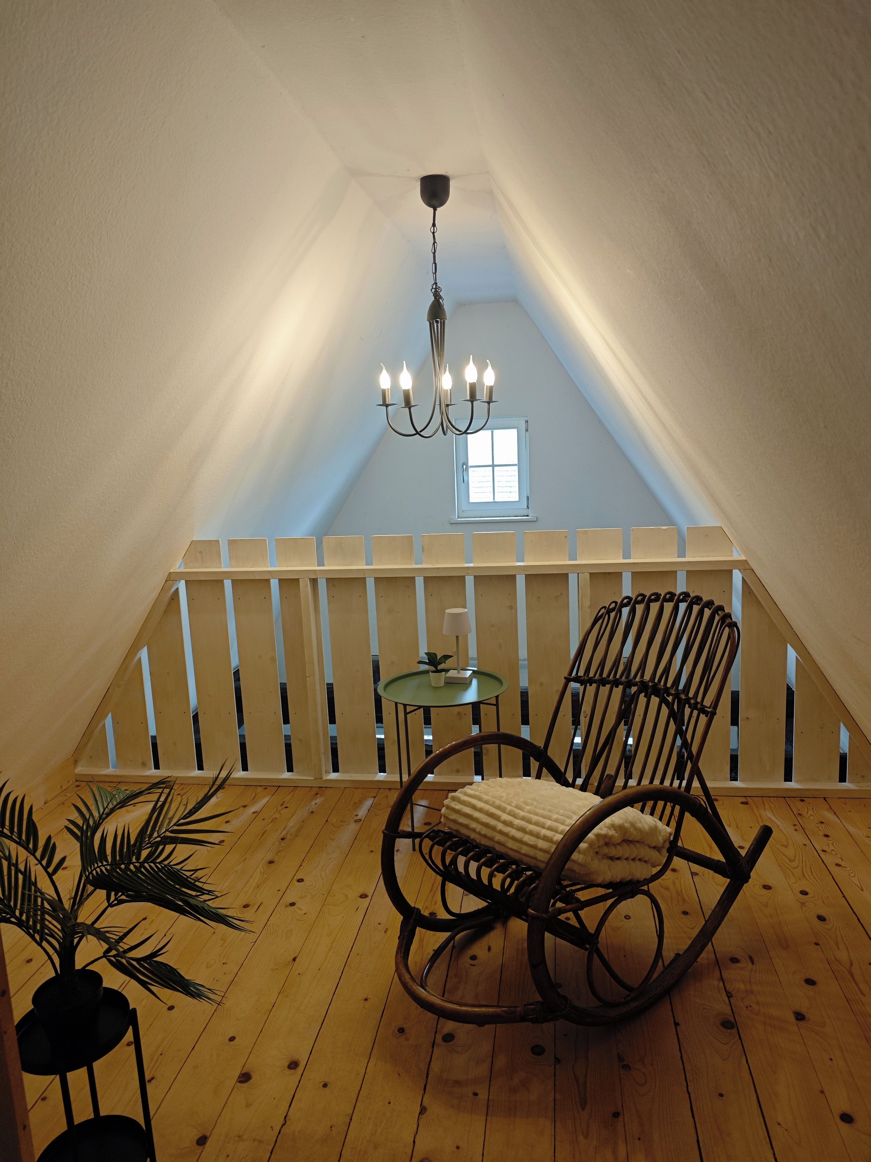 Ein gemütliches Dachzimmer mit schrägen Wänden, einem Holzschaukelstuhl, einem kleinen Tisch mit Lampe und einem Blumentopf.