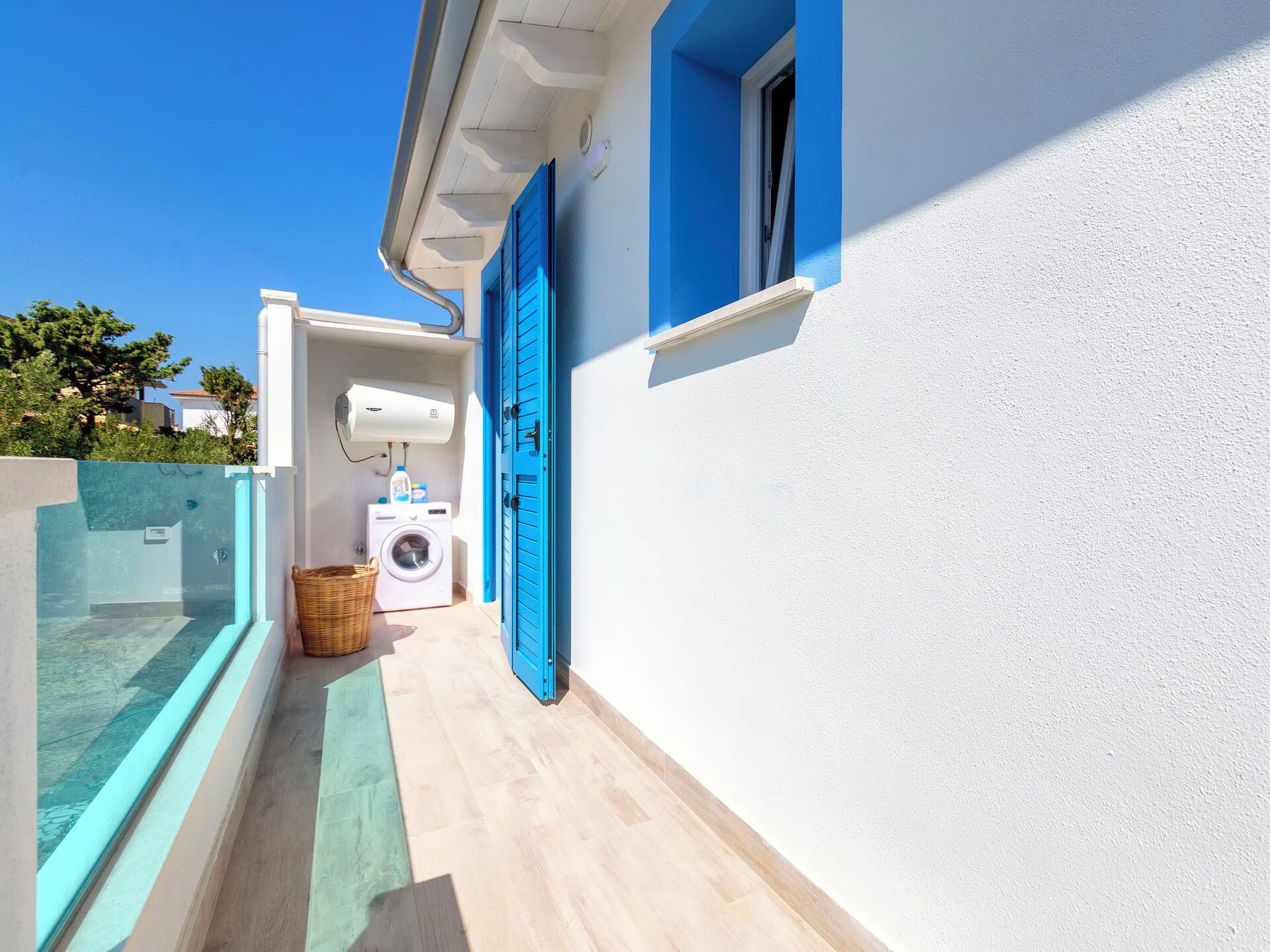 Ein schmaler, überdachter Außenkorridor mit Waschmaschine und Wäschekorb, der zu einem Gebäude mit blauen Fensterläden und weißer Fassade führt.