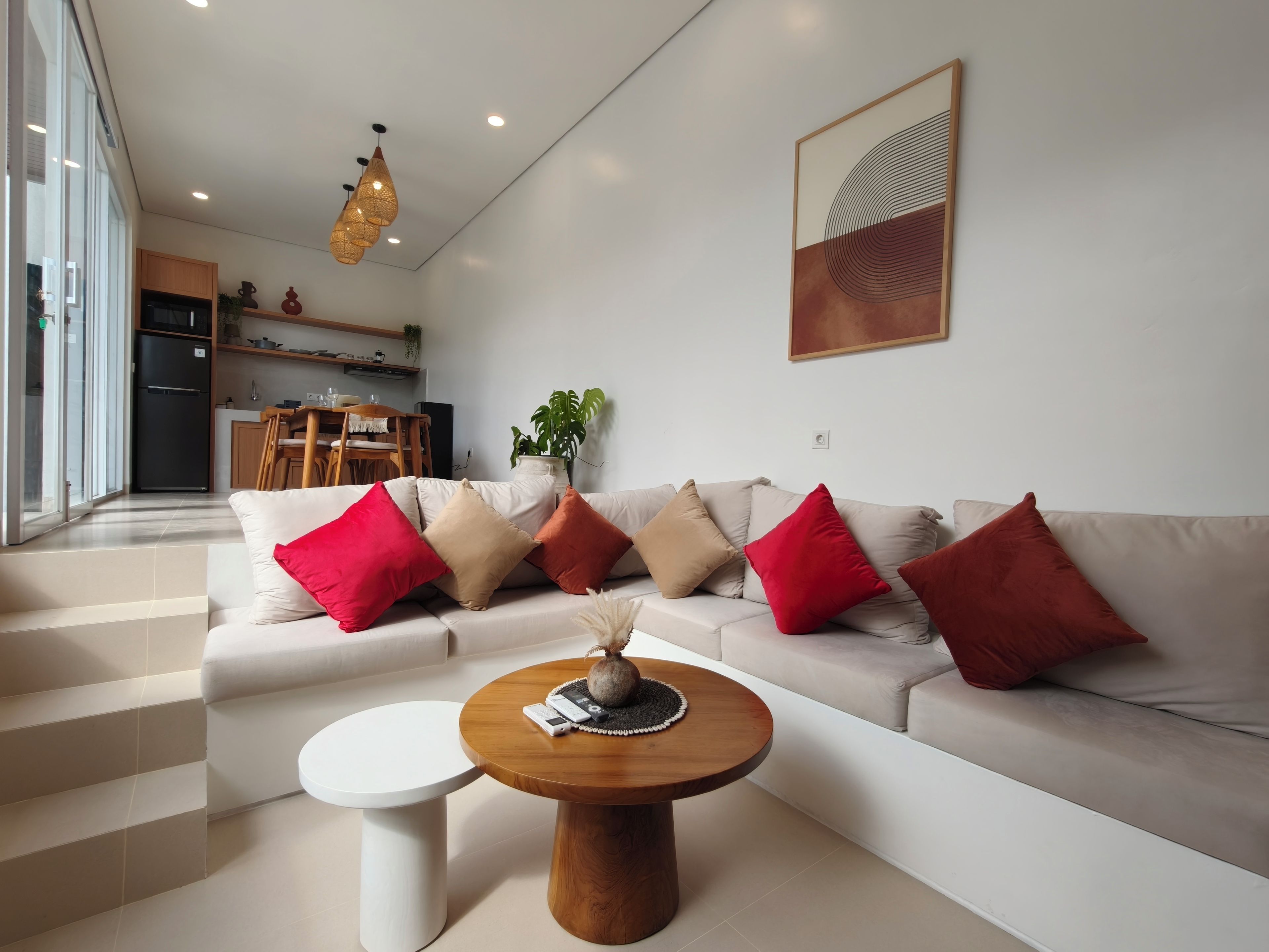 Una sala de estar moderna con un sofá, cojines rojos y beige, una mesa de centro redonda de madera y una mesita auxiliar blanca.