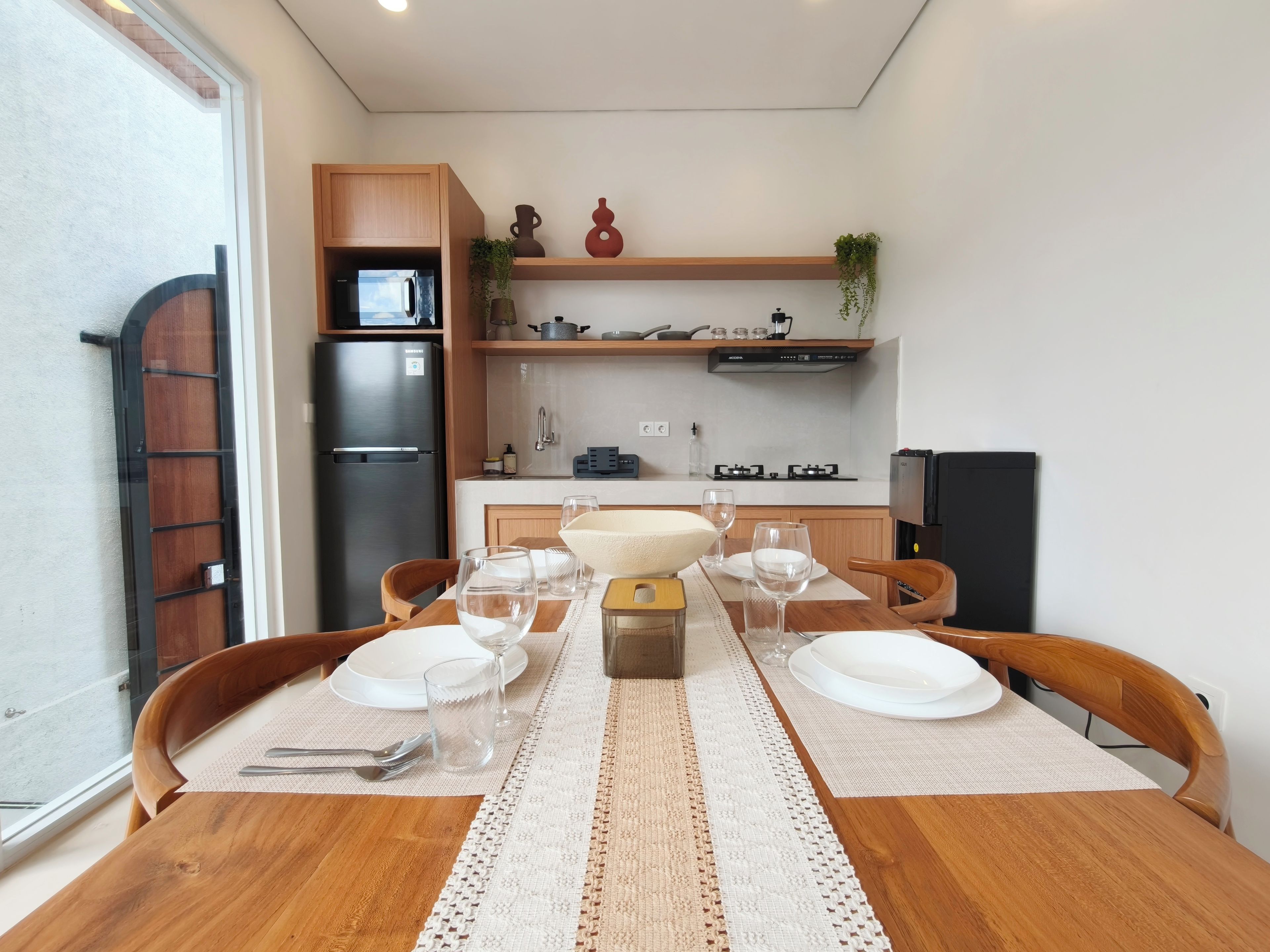 A dining area with a wooden table set for four, a kitchen in the background with appliances and shelves, and a view through a glass door.
