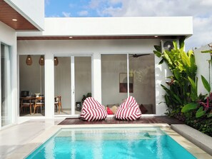 A modern outdoor pool area with a clear blue pool, two striped bean bags, and a dining area visible through glass doors. - Samanaya Villa (Badung)