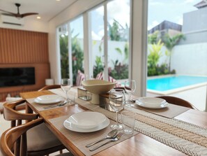 A dining table set with plates, glasses, and cutlery, with a view of a swimming pool and greenery outside. - Samanaya Villa (Badung)