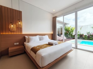 A modern hotel room with a large bed, a wooden headboard, a small bedside table, and a view of a swimming pool through large glass doors. - Samanaya Villa (Badung)