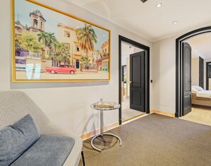 Ein Hotelzimmer mit einem Bett, einem kleinen runden Tisch mit einem Buch, einem grauen Sofa und einem großen Bild in einem Rahmen an der Wand.