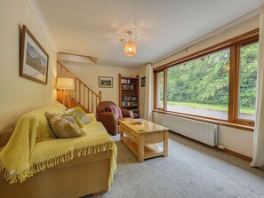 Ein gemütliches Wohnzimmer mit einer Couch, einem Couchtisch, einem Bücherregal und einem großen Fenster mit Blick auf Grünflächen.