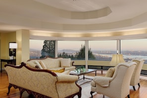 A spacious living room with a large window offering a city view, a classic sofa, and a small table with a lamp.