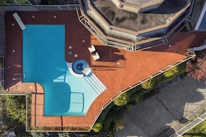 An aerial view of a swimming pool with a surrounding brick deck, a small circular hot tub, and a covered seating area with chairs.