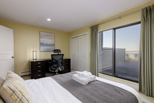 A hotel room with a bed, a desk, a chair, a large window with curtains, and a view of the city.