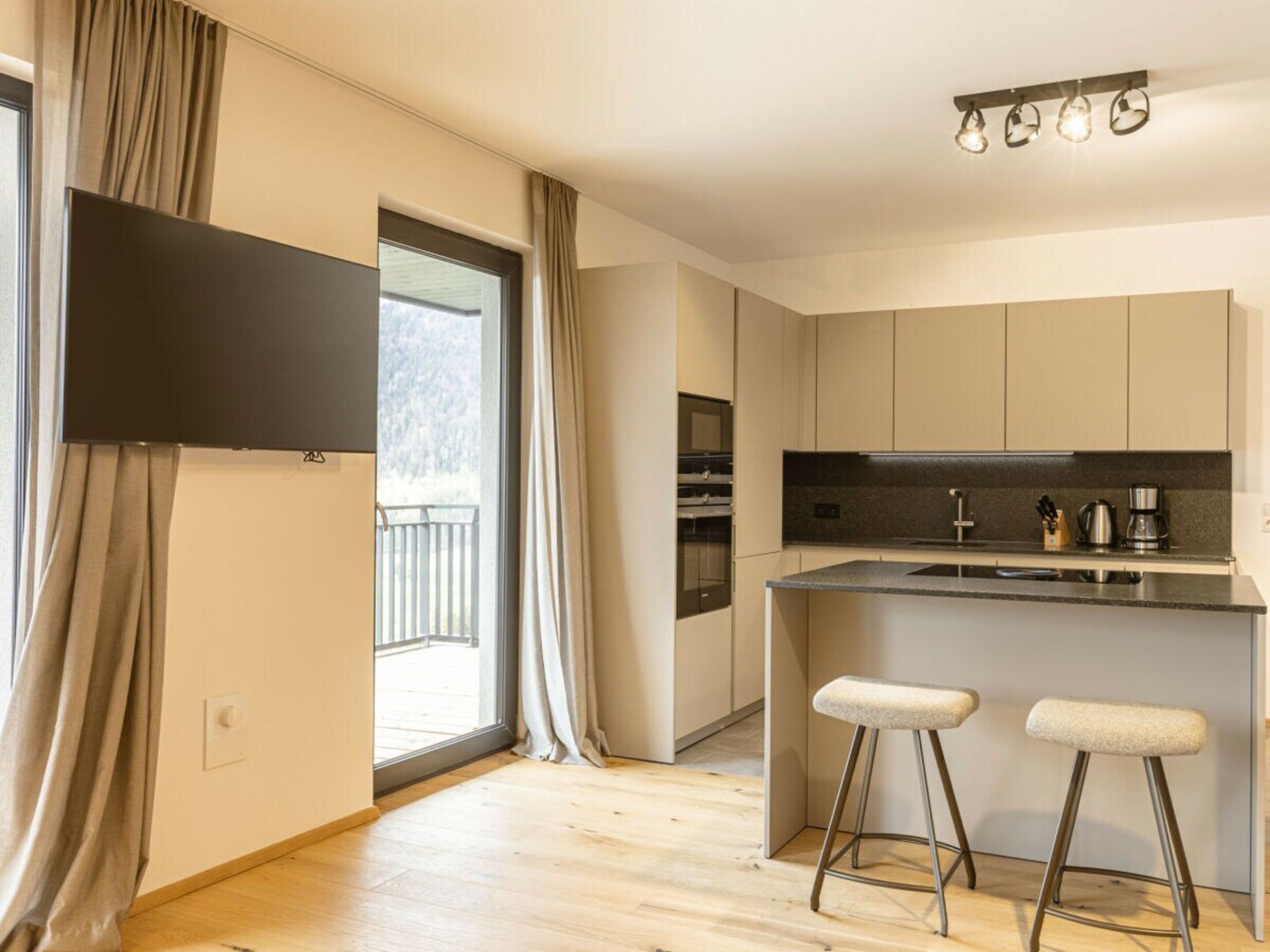 A modern kitchen with a central island, two bar stools, a flat-screen TV, and a window with curtains.