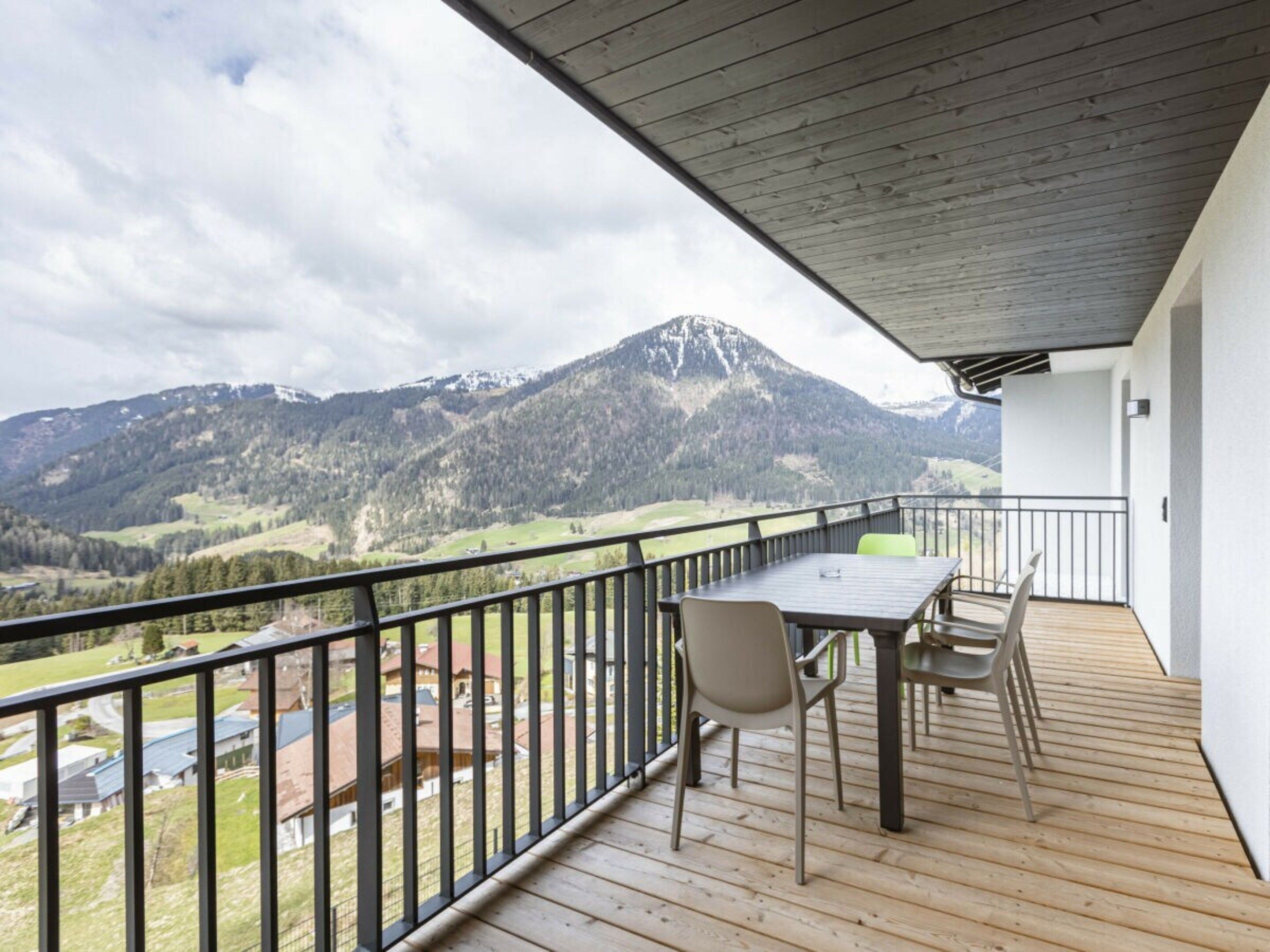 A balcony with a wooden floor, a table, and chairs, overlooking a mountainous landscape.