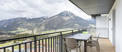 A balcony with a wooden floor, a table, and chairs, overlooking a mountainous landscape.