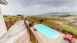 Een houten terras met een zwembad, uitkijkend op een strand en duinen.