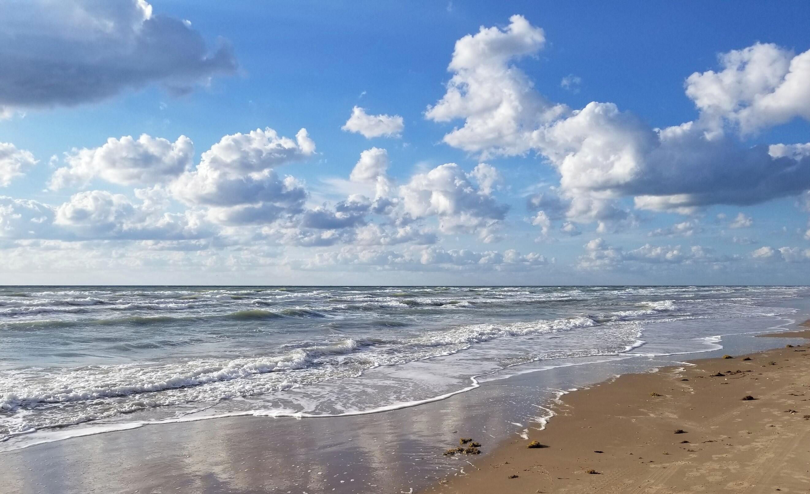 Ein Strand mit Wellen, blauem Himmel und vereinzelten Wolken.