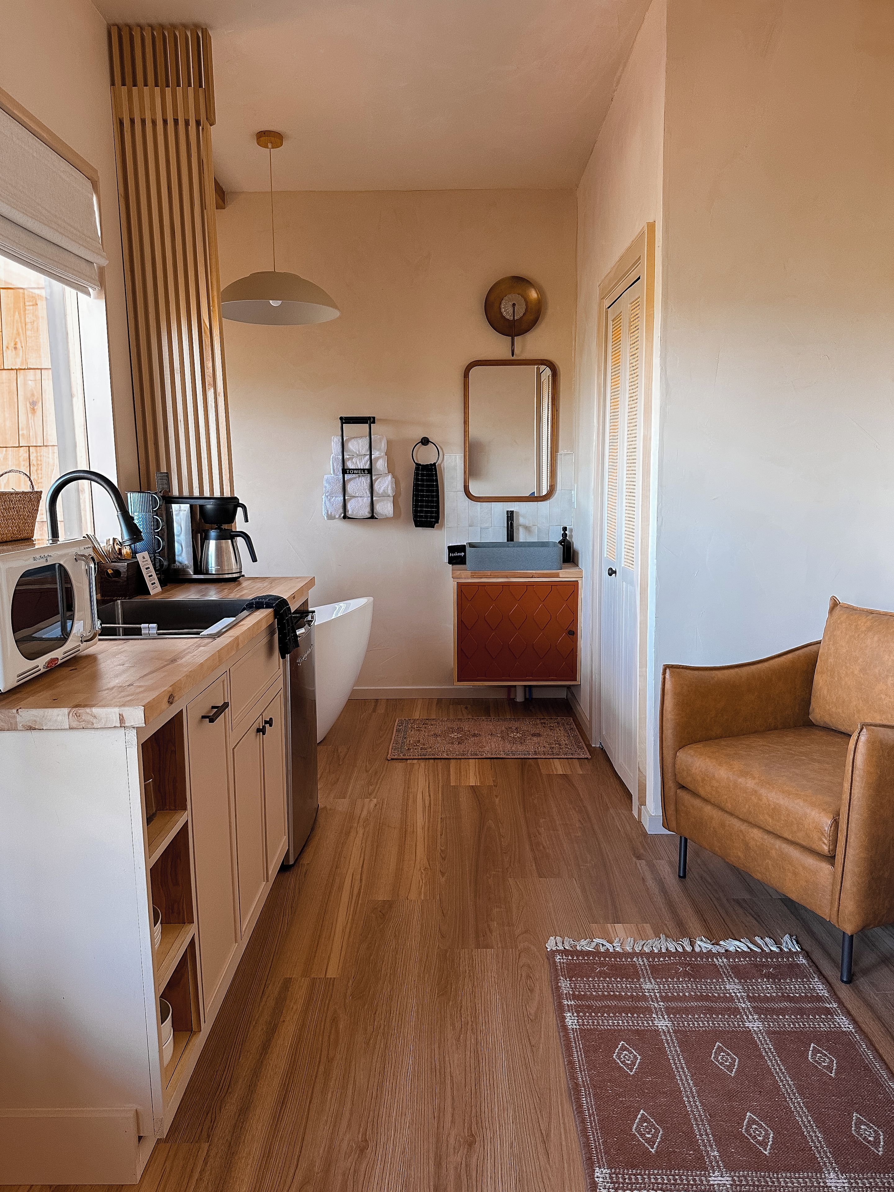 Cocina compacta con microondas, hervidor de agua y cafetera. Se observa un lavamanos con un toallero arriba, y un espejo en la pared. Una silla de cuero marrón es visible en la esquina.