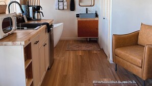 Cozinha compacta com micro-ondas, chaleira e máquina de café. Há uma pia com um porta-toalhas acima e um espelho na parede. Uma poltrona de couro marrom é visível no canto.