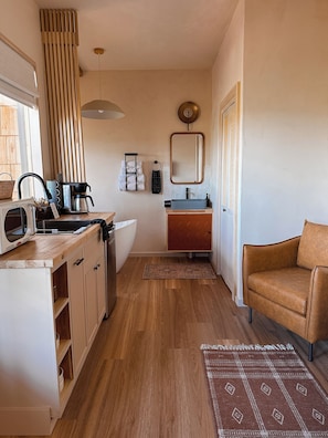 A compact kitchen with a microwave, kettle, and coffee maker. There is a sink with a towel rack above it, and a mirror on the wall. A brown leather chair is visible in the corner. - The Canopy · Treetop Cabin, Hot Tub + Soaking Tub (Valley Mills)