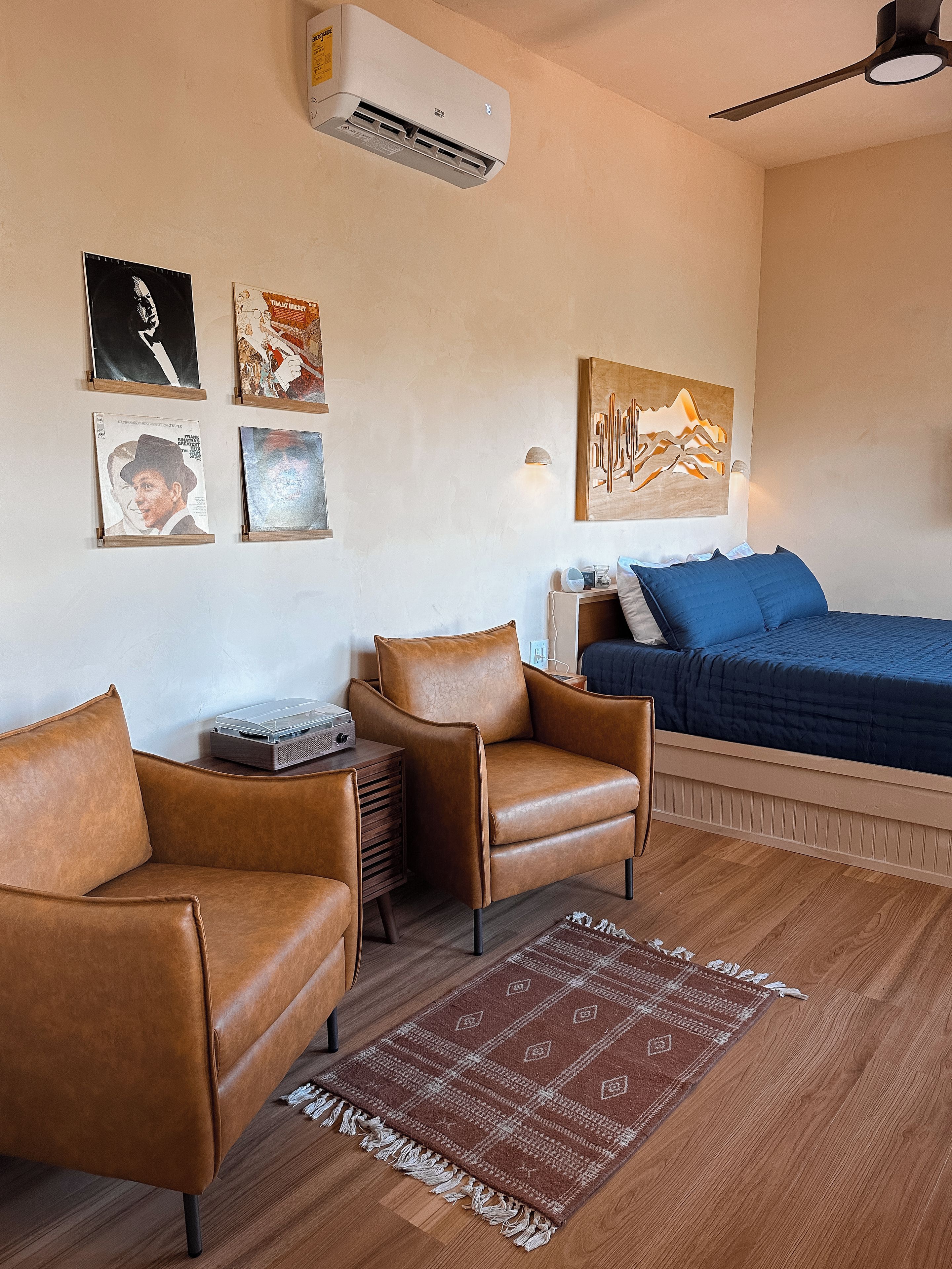 A room with a bed, two leather armchairs, a small table with a radio, a wall-mounted air conditioner, and a rug.