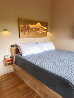 A neatly made bed with a quilted grey cover, a bedside table with a lamp, and a wall-mounted artwork depicting a desert landscape. - Treetop Cabin: Hot Tub, Outdoor Shower & Views (Valley Mills)