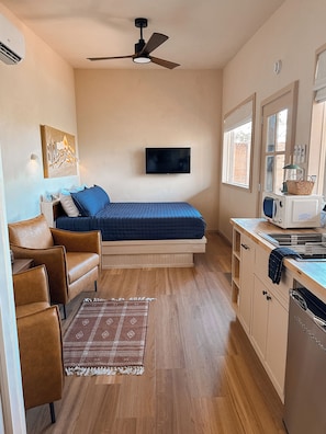 A compact room with a bed, a sofa, a television, a microwave, and a small kitchen area. - The Canopy · Treetop Cabin, Hot Tub + Soaking Tub (Valley Mills)