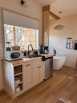 A modern kitchen with a built-in dishwasher, a large window with blinds, a wooden ceiling, and a white sink. - The Canopy · Treetop Cabin, Hot Tub + Soaking Tub (Valley Mills)