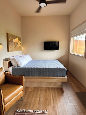 A bedroom with a bed, a TV mounted on the wall, a ceiling fan, and a window with a view of a wooden fence.