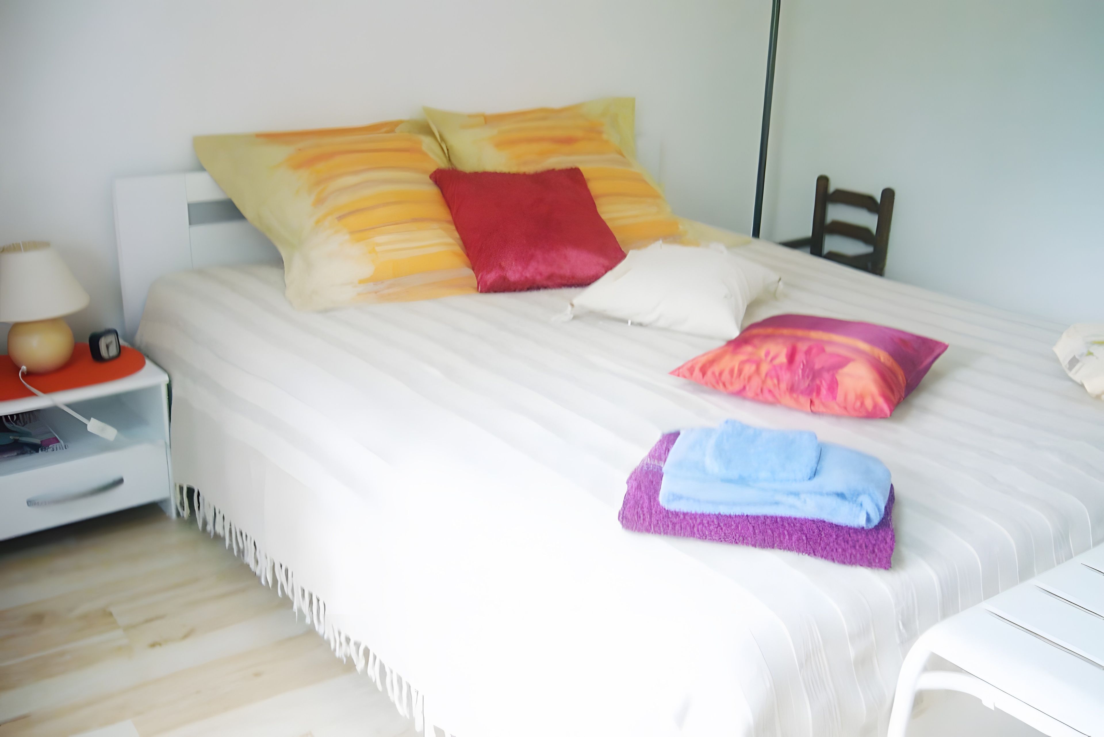 A neatly made bed with multiple pillows, a bedside table with a lamp and a clock, and a chair in the corner.