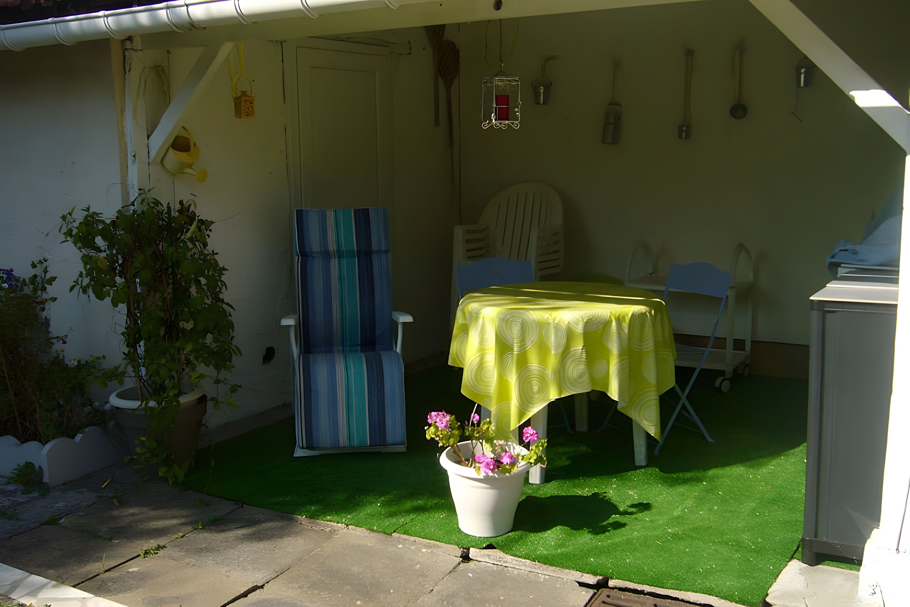 Eine Terrasse mit einem Tisch, der mit einer gelben Tischdecke bedeckt ist, einem blauen Stuhl, einem Topf mit rosa Blumen und einem grünen Rasen aus Kunstfaser.
