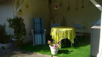 Eine Terrasse mit einem Tisch, der mit einer gelben Tischdecke bedeckt ist, einem blauen Stuhl, einem Topf mit rosa Blumen und einem grünen Rasen aus Kunstfaser.
