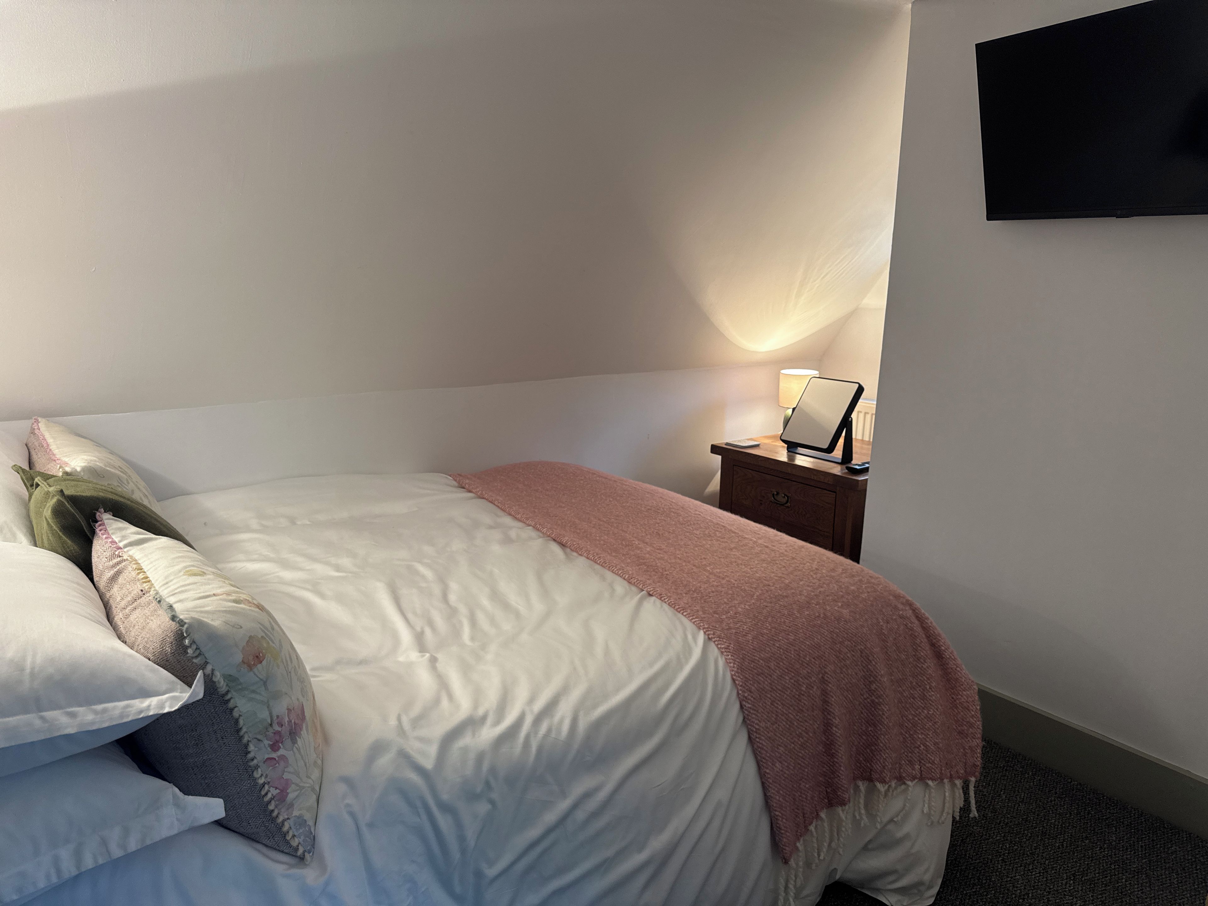 A neatly made bed with pillows, a bedside table with a lamp, and a flat-screen TV on the wall.