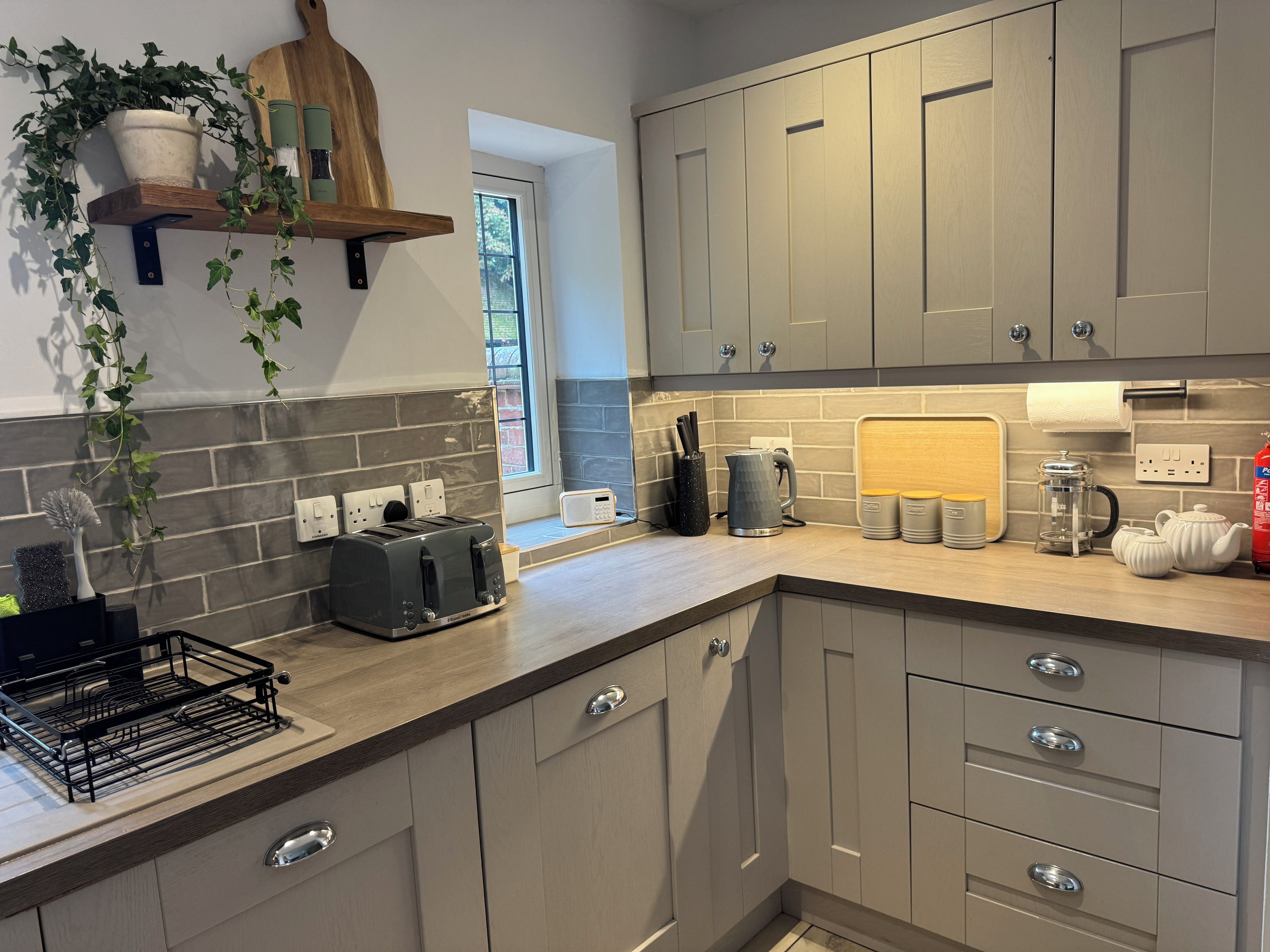 A modern kitchen with grey cabinets, a wooden shelf with a potted plant, a toaster, a coffee maker, and a kettle on the counter.