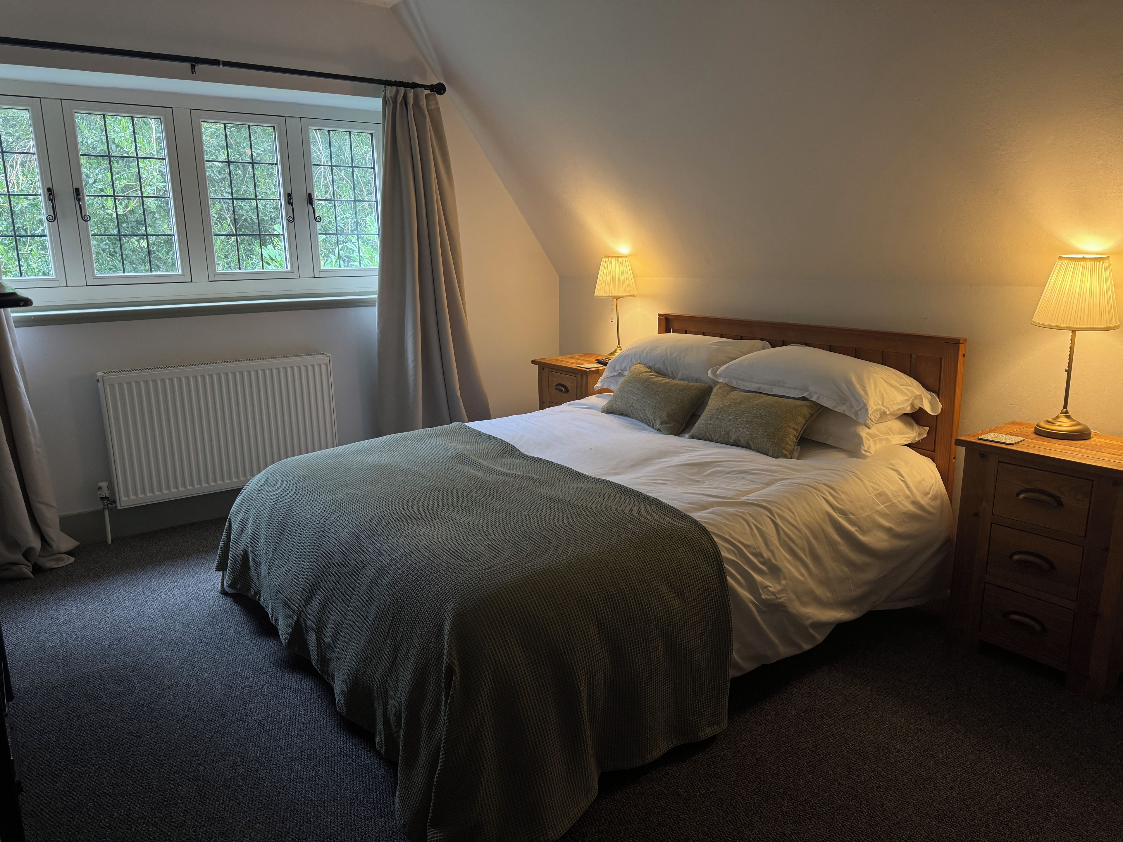 A bedroom with a bed, two bedside tables, a window with curtains, a radiator, and a lamp on each bedside table.