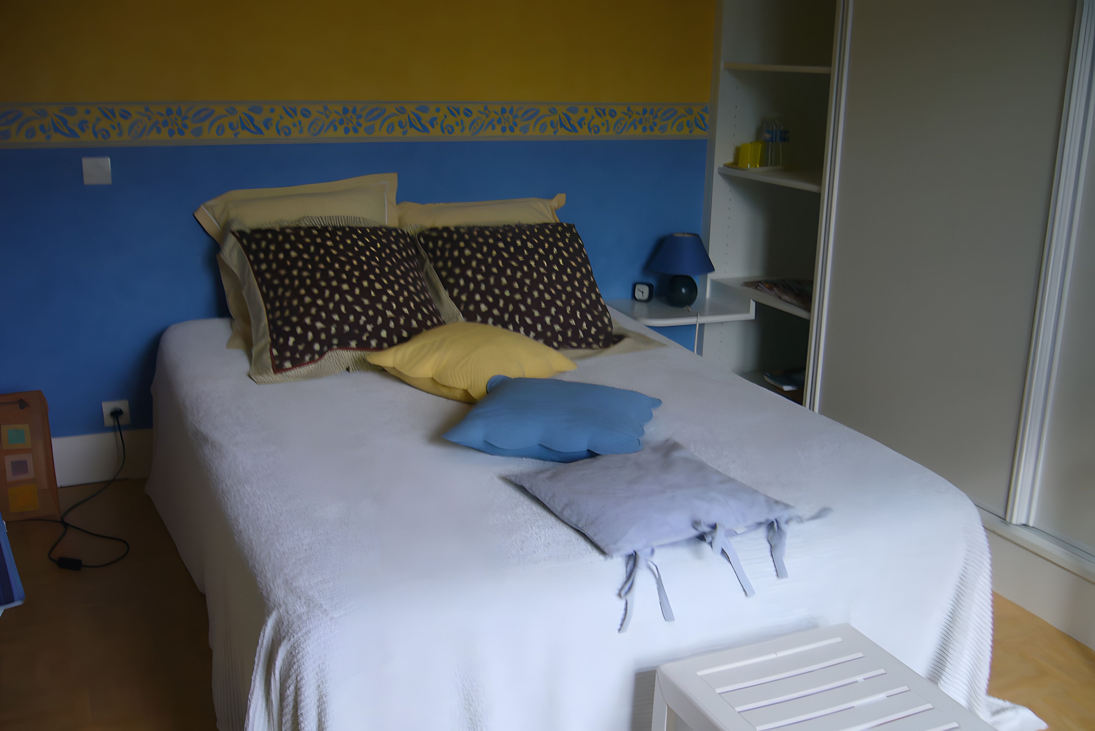 A bed with multiple pillows, a bedside table with a lamp, and a power outlet on the wall.