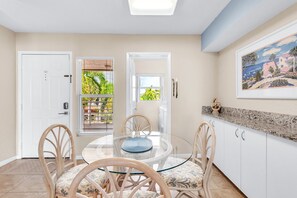 A dining area with a round glass table and four chairs, a white door, a window with blinds, and a painting on the wall. - Resort Style Amenities + Quick Drive to Beach | Bahia Vista 478 (Saint Petersburg)