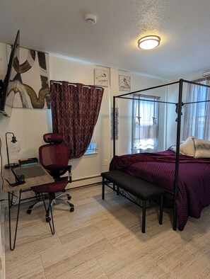 A bedroom with a four-poster bed, a desk with a chair, a window with blinds, and a ceiling light. - 146-4 Miles Davis, Jazz Great! A place 2 Rest & Relax.  	1 Bedroom Studio. (New London)