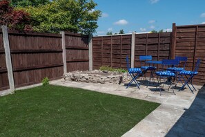 Ein Garten mit Holzzaun, Steinmauer und Rasen. Blaue Klappstühle und ein Tisch sind aufgestellt.